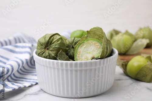 Fresh green tomatillos with husk in bowl on light marble table, closeup photo