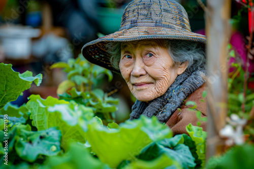 Old woman wearing hat and scarf in garden filled with green plants.
