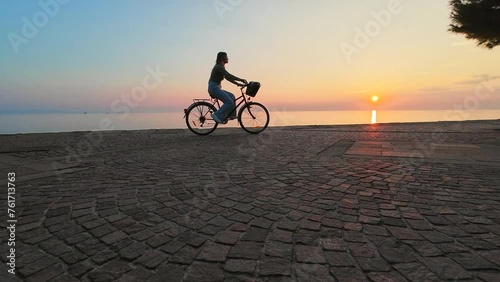 Wide tracking shot of a young woman cycling on a coastal path as the sun is setting in the background.