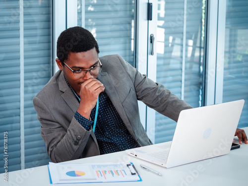 A handsome African American businessman in a suit is sitting at a work desk analyzing financial data and doing research