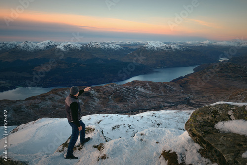 A hiker on top of a mountain looks out over the magnificent scenery of a lake surrounded by mountains in winter. Loch Lomond and The Trossachs National Park. Scotland