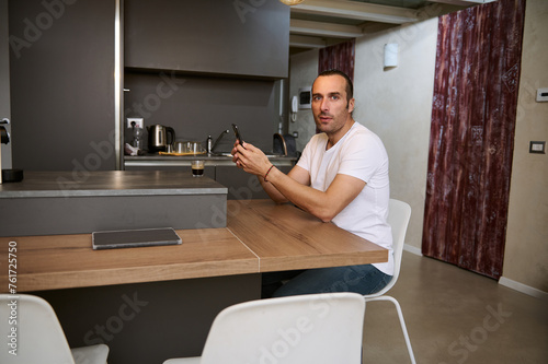 Smiling young man looking at camera while using smartphone for online shopping  sitting at table at home