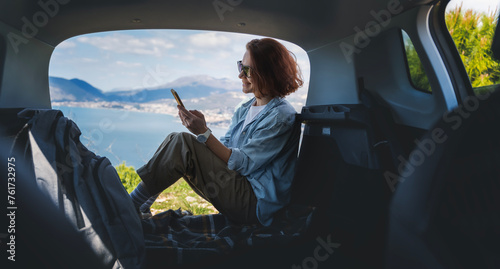 Young woman sitting in the open trunk of a car overlooking the sea with a smartphone in her hands, summer vacation and auto travel © olezzo