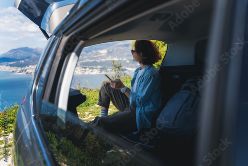 Young woman sitting in the open trunk of a car overlooking the sea with a smartphone in her hands, summer vacation and auto travel