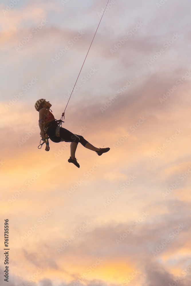 A woman is hanging from a rope, and the sky is orange and pink. Scene is adventurous and exciting A woman is hanging from a rope, and the sky is orange and pink. Scene is adventurous and exciting