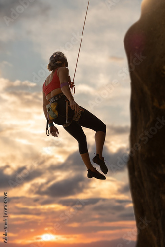 A woman is climbing a rock wall with a rope. The sky is orange and the sun is setting