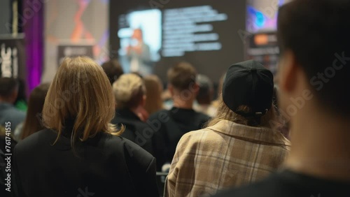 At the frontier of innovation, a presenter articulates the nuances of emerging technologies to an auditorium filled with aspiring entrepreneurs. In foreground there is blonde and girl in cap.