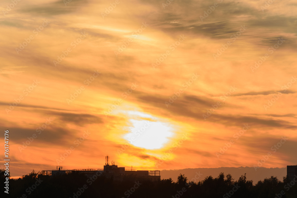 The setting sun in the yellow-orange evening sky over the black silhouette of city buildings on the horizon