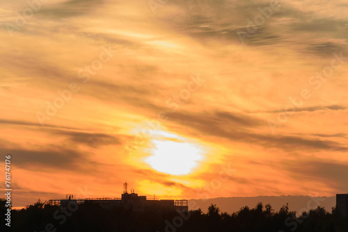 The setting sun in the yellow-orange evening sky over the black silhouette of city buildings on the horizon