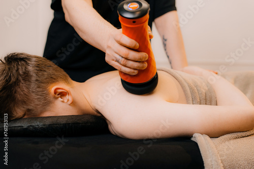 Therapist massaging a man's back with a percussion massage device in a massage room. The therapist's hand holds a therapeutic vibratory massager. Physiotherapy and Muscle Recovery and Massage