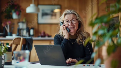 Senior business woman talking on the phone while working in her own office, laptop standing on the table, woman looking into the frame, senior generation experience and knowledge
