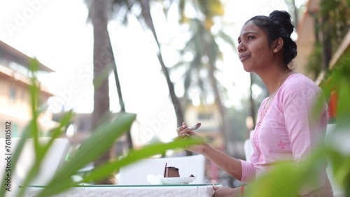 Pretty young woman with dark skin enjoys sweet dessert, sits at table in afternoon on street, student of Indian nationality. Pleasant moments of rest. Breakfast in fresh air.