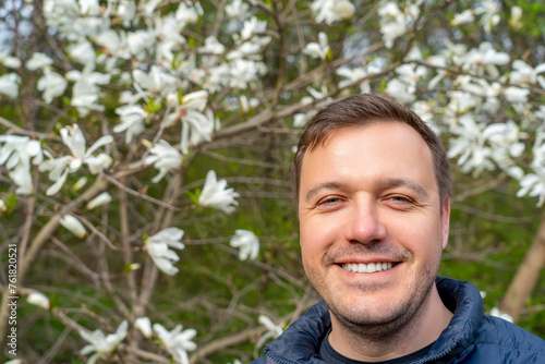 A cheerful man in a jacket stands in focus against a backdrop of vibrant white blossoms under a bright sky