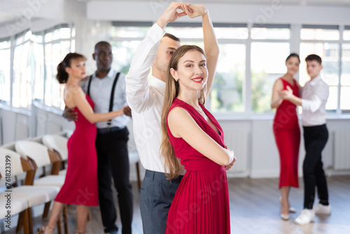 Happy young woman dressed in red dress with man in elegant clothes dancing bachata in modern ballroom dance class