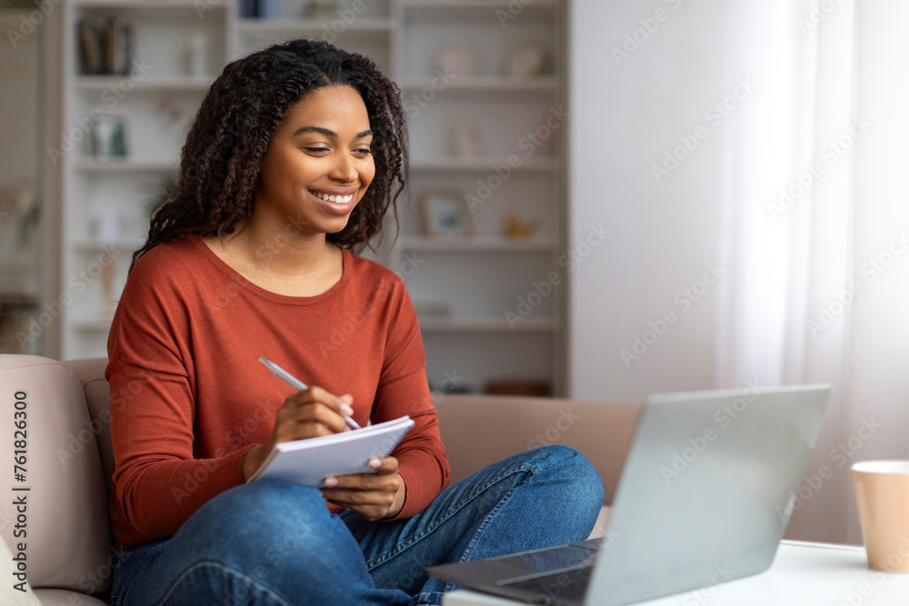 Smiling young black woman writing down notes from her laptop session ...