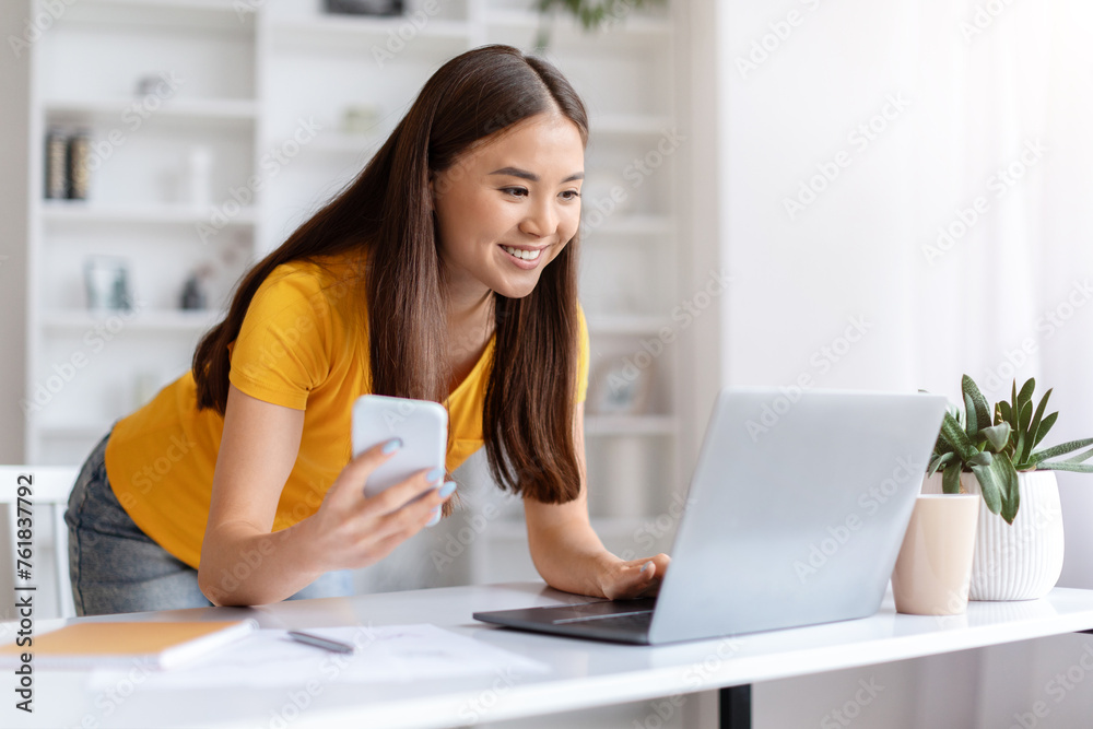 Cheerful asian woman using smartphone while leaning towards her laptop at home Cheerful asian woman using smartphone while leaning towards her laptop at home
