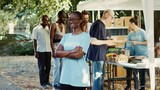 Side-view of black woman wearing glasses outside with arms crossed and eyes fixed on camera. Multiethnic volunteers supporting nonprofit initiative aimed at reducing hunger and aiding those in need.