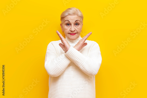 old grandmother in a white sweater shows a stop gesture with her arms crossed and prohibits on a yellow isolated background, elderly woman pensioner shows refusal and prohibition photo