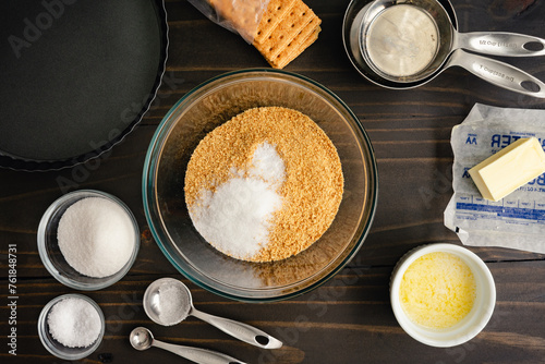 Graham Cracker Crust Ingredients with a Tart Pan: Graham cracker crumbs and sugar in a mixing bowl surrounded with other ingredients and kitchen tools