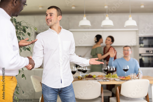 Genial young man in casual attire extending warm handshake to friend, welcoming to enjoyable home dinner event