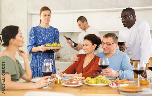 Cheerful dinner scene with multiethnic adult friends toasting with wine  enjoying snacks  and engaging in lively conversations in cozy home kitchen ..