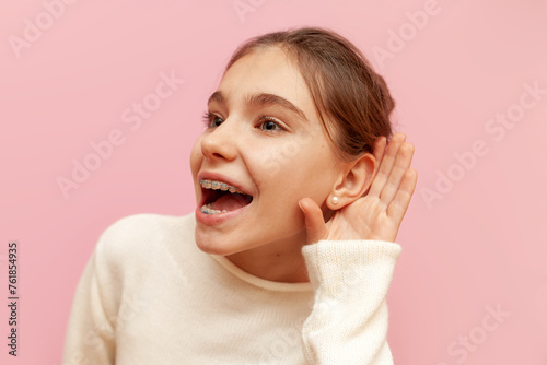 curious teenage girl with braces eavesdrops on secret information on a pink isolated background, child holds hand near ear and listens