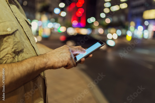 Close up View of Smart Phone in Male Hand in the City Streets with Illuminated Lights in Background