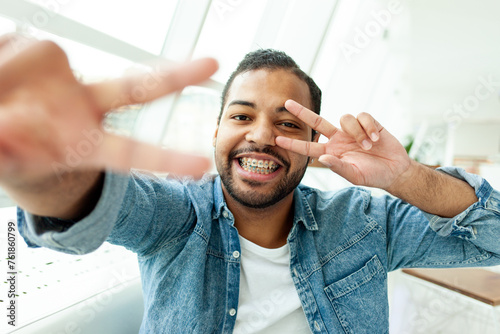 cherful African American man with braces smiles and shows peace gesture in white room, man with stubble shows two fingers and greets photo