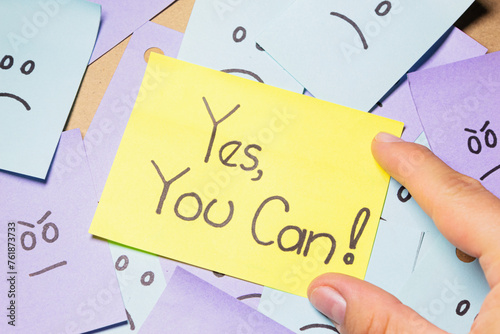 A person holds a sticky note with the message ¨Yes, you can!¨ over a pile of sticky notes with sad and angry faces. Motivational message. photo