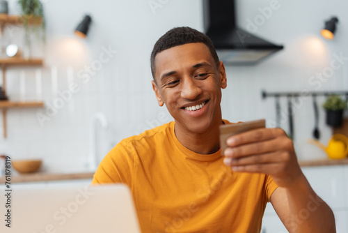 Happy African American man holding credit card, using laptop, choosing, paying, online shopping