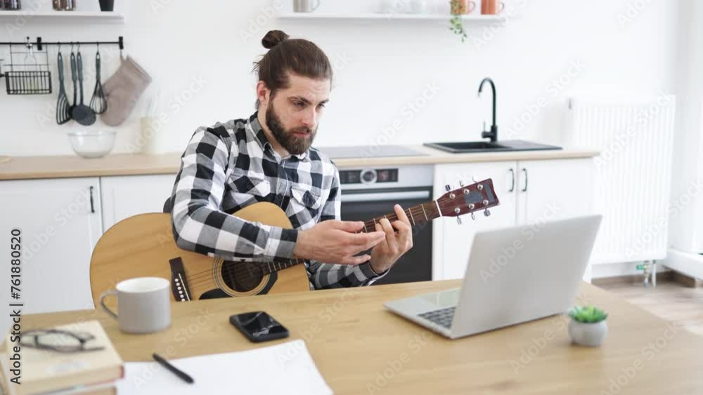 Cheerful Caucasian person holding string instrument while smiling at portable computer webcam in spacious apartment. Happy male teaching online guitar lesson using modern technologies. Cheerful Caucasian person holding string instrument while smiling at portable computer webcam in spacious apartment. Happy male teaching online guitar lesson using modern technologies.