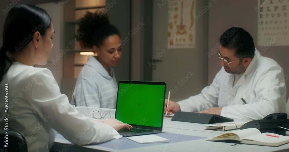 The nurse uses a laptop with a green screen, entering patient data. A woman talks to a doctor sitting at a table in the background