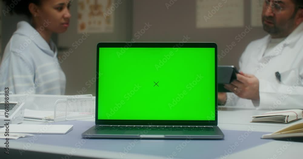 A laptop with a green screen is on the table in the medical office. A doctor is talking to a female patient sitting in the background A laptop with a green screen is on the table in the medical office. A doctor is talking to a female patient sitting in the background