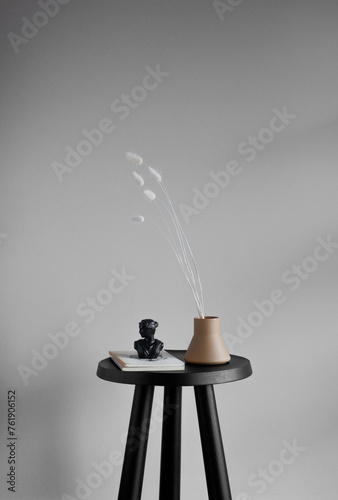 Still life of vintage books, black sculpture bust, and ceramic vase with white Foxtail stems on a small black table