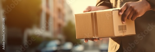The hands of the delivery man carry the package to deliver. Delivery man's hand holding brown box, transport truck background Detail of a delivery man holding a labeled cardboard package.