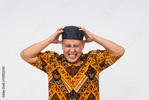 Seething Indonesian man in batik shirt and kopiah expressing frustration, clutching his head. isolated on a white background with copy space. © Mdv Edwards