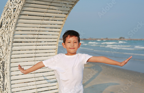 Portrait of smiling Asian boy open arms wide while standing on white bamboo wooden sofa bed with looking camera at the beach.