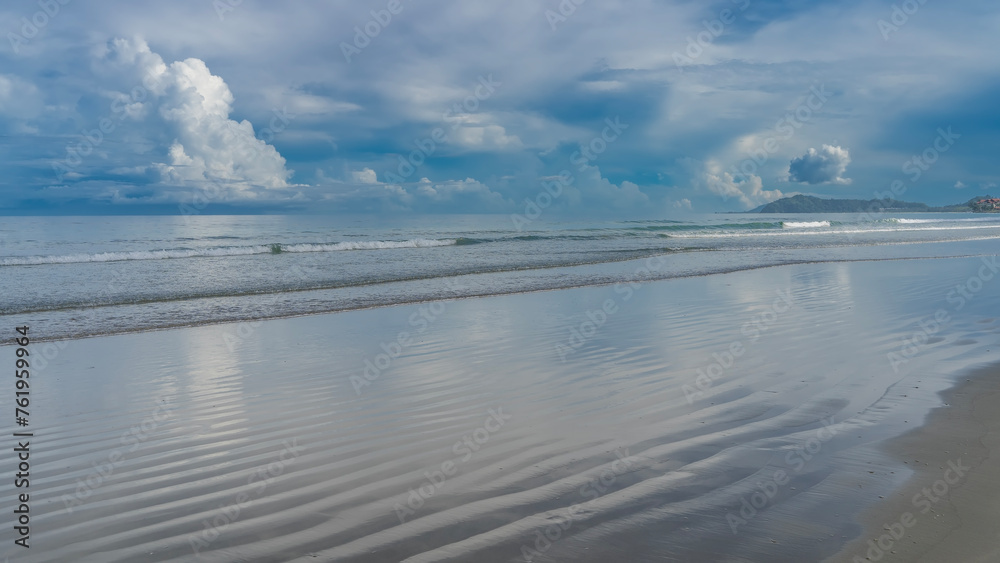 The waves of the turquoise ocean are calmly rolling towards the shore, spreading along the beach. The blue sky and clouds are reflected on the wet wavy sand. Mountains on the horizon. Malaysia. Borneo