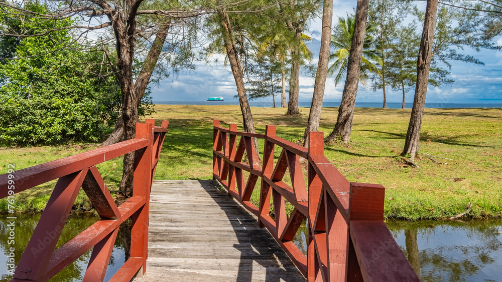 A wooden pedestrian bridge with railings spans over a small river. Bushes and palm trees grow on the green lawn. There's an ocean ahead. A ship on the horizon against a blue sky and clouds. Malaysia A wooden pedestrian bridge with railings spans over a small river. Bushes and palm trees grow on the green lawn. There's an ocean ahead. A ship on the horizon against a blue sky and clouds. Malaysia