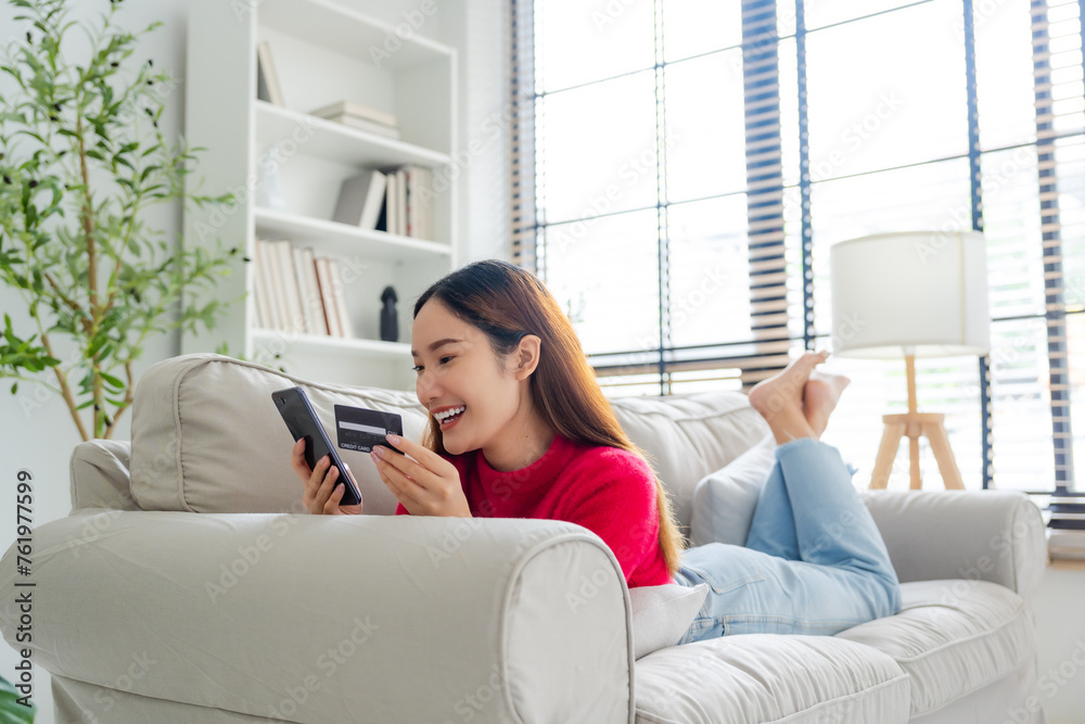 Young asian woman lying on sofa in living room, makes online banking payments through the internet from bank card on cell phone. Shopping online on mobile with credit card Young asian woman lying on sofa in living room, makes online banking payments through the internet from bank card on cell phone. Shopping online on mobile with credit card