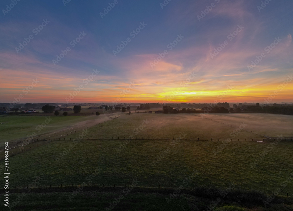 This wide-angle photograph captures the tranquil beauty of a sunrise spreading its warm glow over a lush pasture. The horizon is adorned with an array of colors, ranging from deep purples to fiery