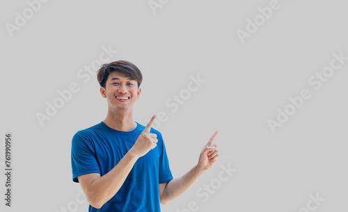 A young Asian man in his 20s wearing a blue t-shirt happy smiling pointing thumbs up isolated on a gray background. The person's index finger pointing to a blank space for text. Positive person.