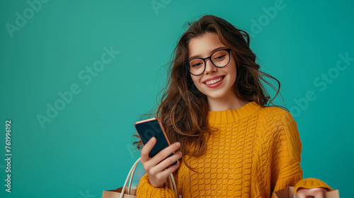 Portrait of a happy young woman holding shopping bags and using mobile phone on teal color background professional photography.