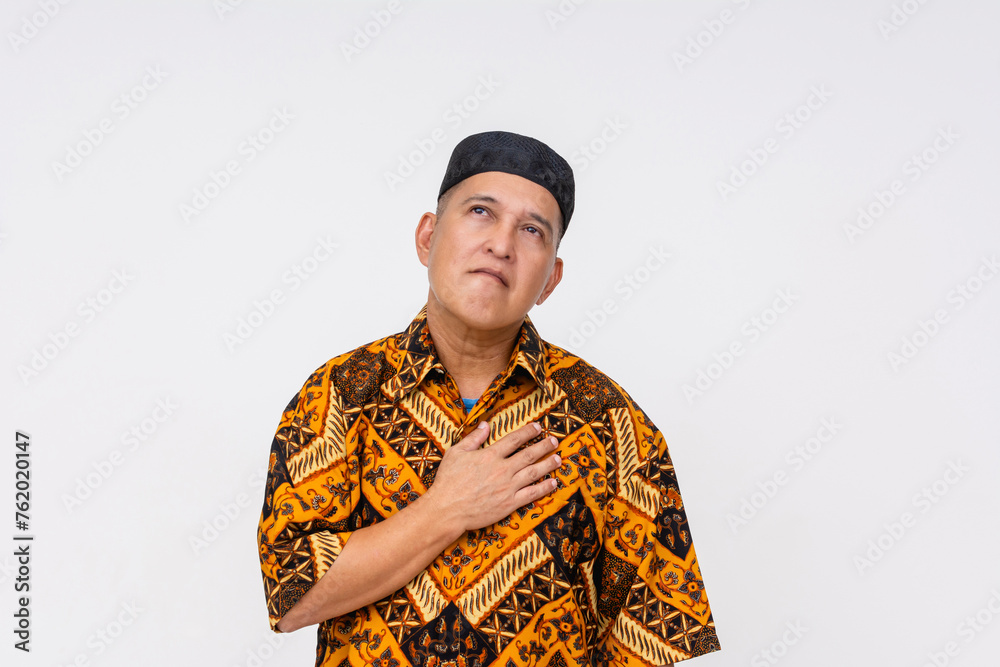 Indonesian man in traditional batik attire and kopiah hat looking up experiencing chest tightness, isolated on a white background. Indonesian man in traditional batik attire and kopiah hat looking up experiencing chest tightness, isolated on a white background.