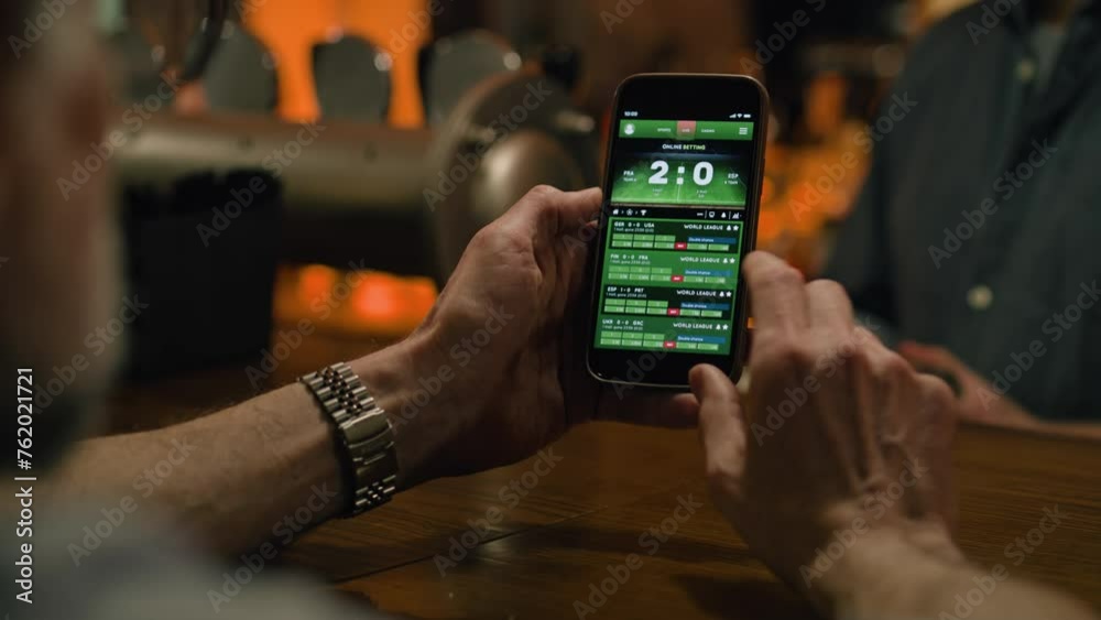 Close up of mature sports fan checking game score and bookmaker ratings in app using mobile phone. Caucasian man bets on football match online sitting at bar counter in pub at night. Gambling concept.
