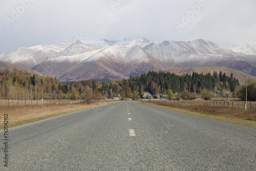 Long rural road leading to the mountains in Twizel, New Zealand. Snow-capped mountains can be seen in the background with a forest at the mountain foothills. 