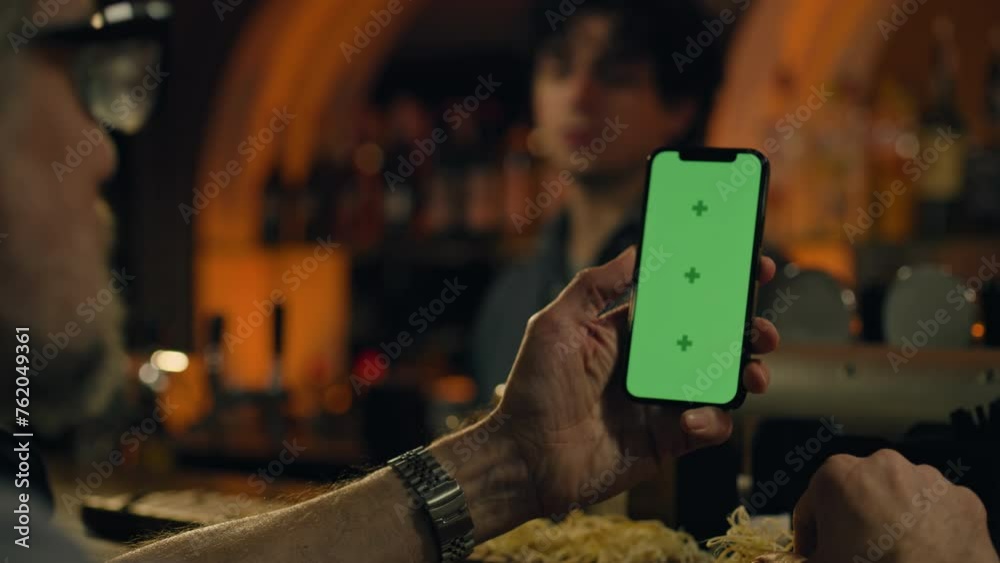 Mature man uses mobile phone with green screen, bets on football match online or surfs the Internet. Sports fan checks bookmaker ratings, talks with bartender sitting at bar counter in pub. Chroma key