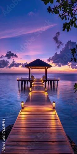 A long wooden pier with lights leads to an overwater gazebo at dusk.