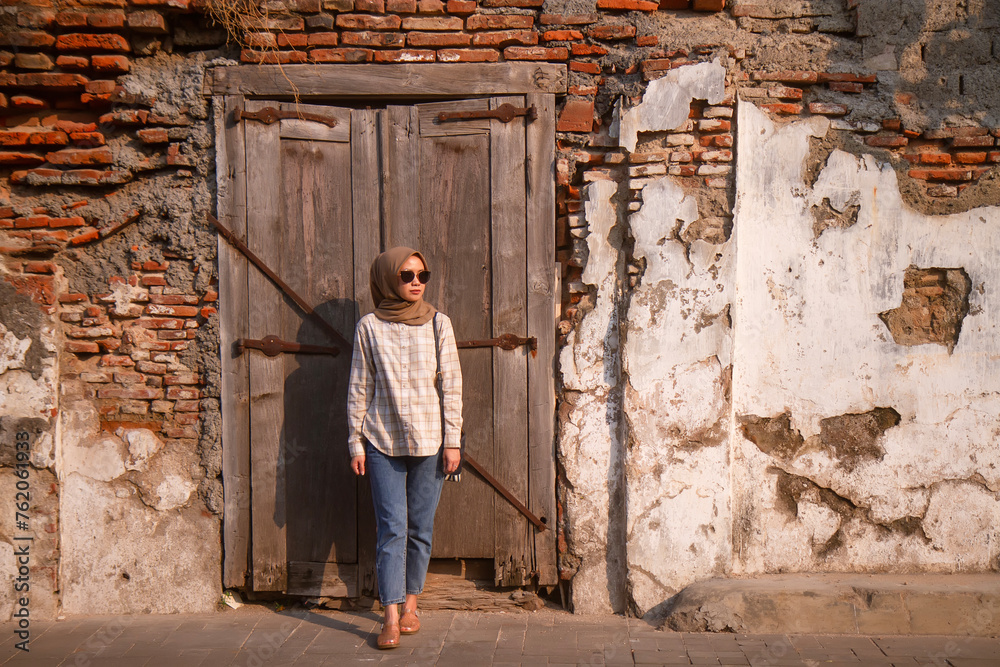 happy asian muslim woman wearing sunglasses standing in front of wooden door with old grunge brick wall while roaming around kota lama or old city area, Semarang, Central Java , Indonesia