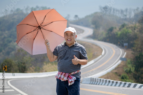 Asian middle-aged man in white cap and wears loincloth, holding umbrella and standing beside the road like number 3 on high mountain in Nan, province, northern part of thailand, soft focus. photo